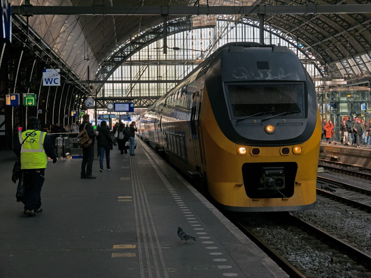 A yellow NS Intercity train at a platform under the glass roof of Amsterdam Centraal, passengers walking alongside